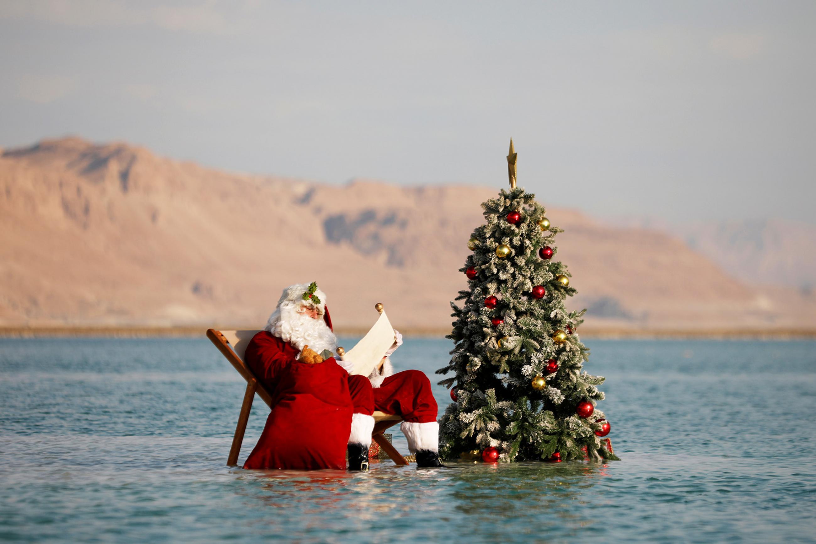 A man, wearing a red Santa Claus costume complete with a hat, beard, and bag full of toys, holds a scroll as he poses for a picture while sitting next to a decorated Christmas tree on a salt formation in the Dead Sea, near Ein Bokeq, Israel on November 15, 2020.