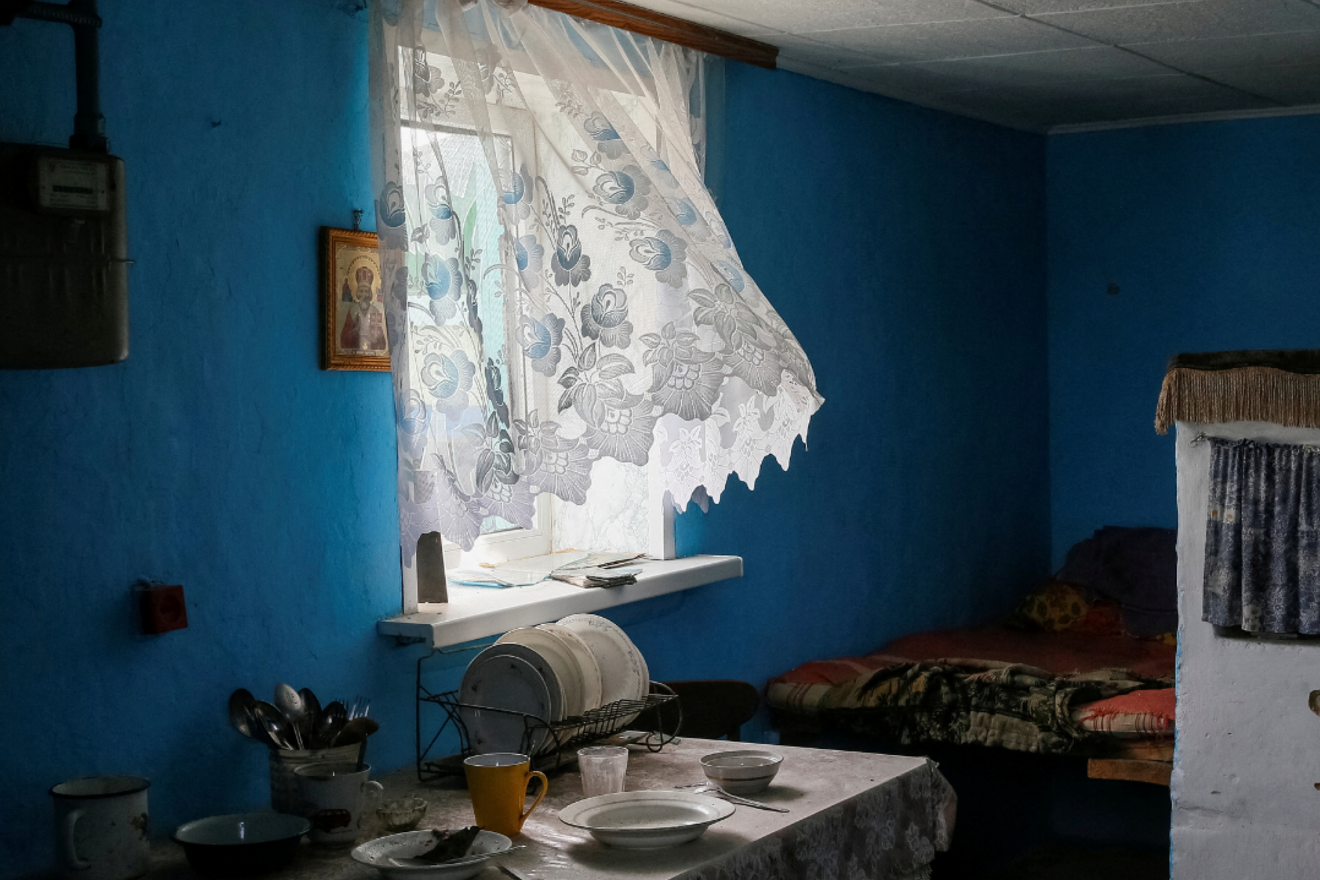 A view of a corner of the kitchen with cheerfully painted blue walls, dishes left on the counter, and white lace curtain fluttering in the wind — abandoned by residents amid the Russian war on Ukraine. The Kyiv region, March 2022.