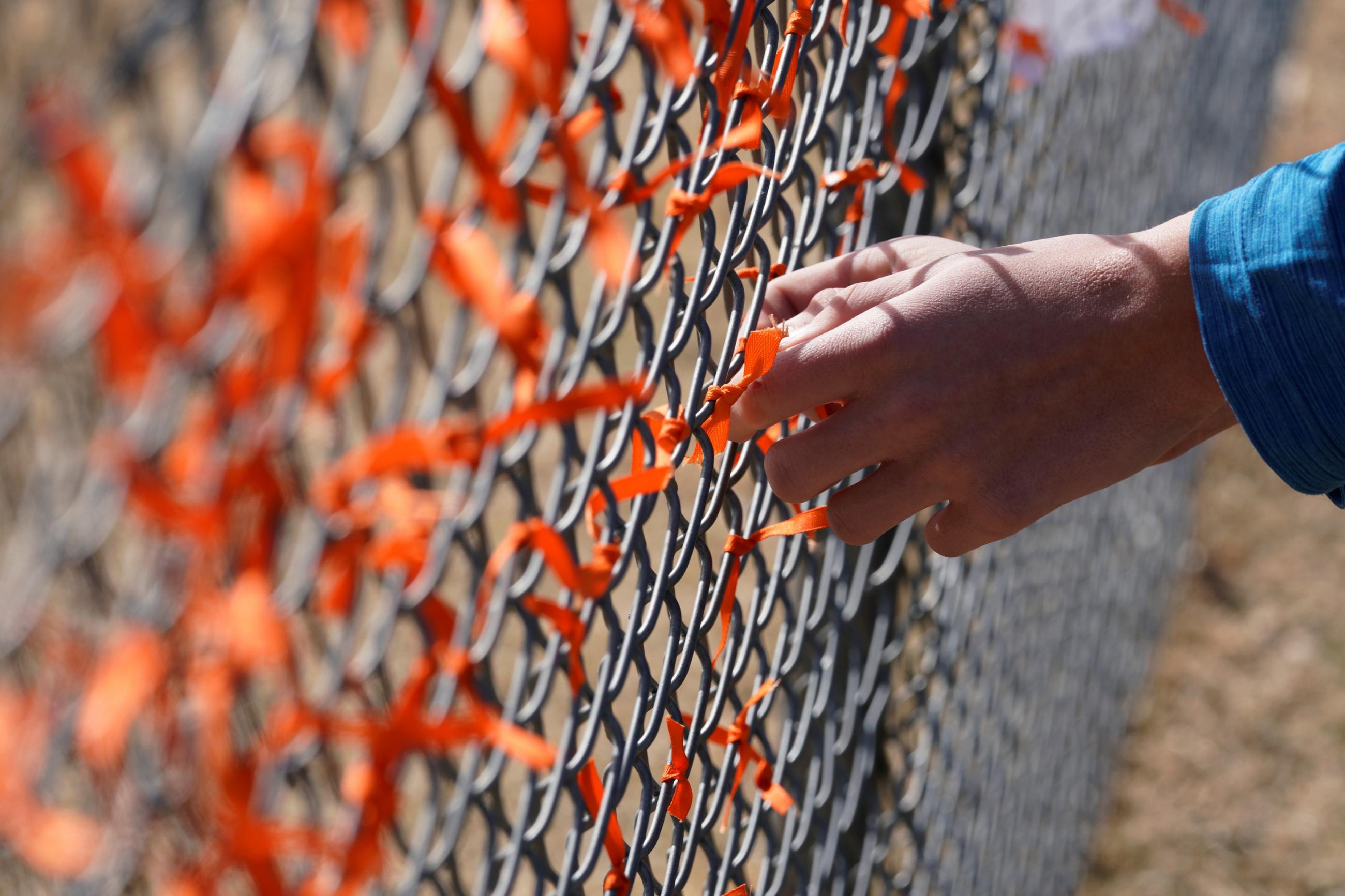 A white hand with the cuff of a blue jacket reaches out to tie an orange ribbon onto a chain-link fence covered in orange ribbons in honor of those killed by gun violence