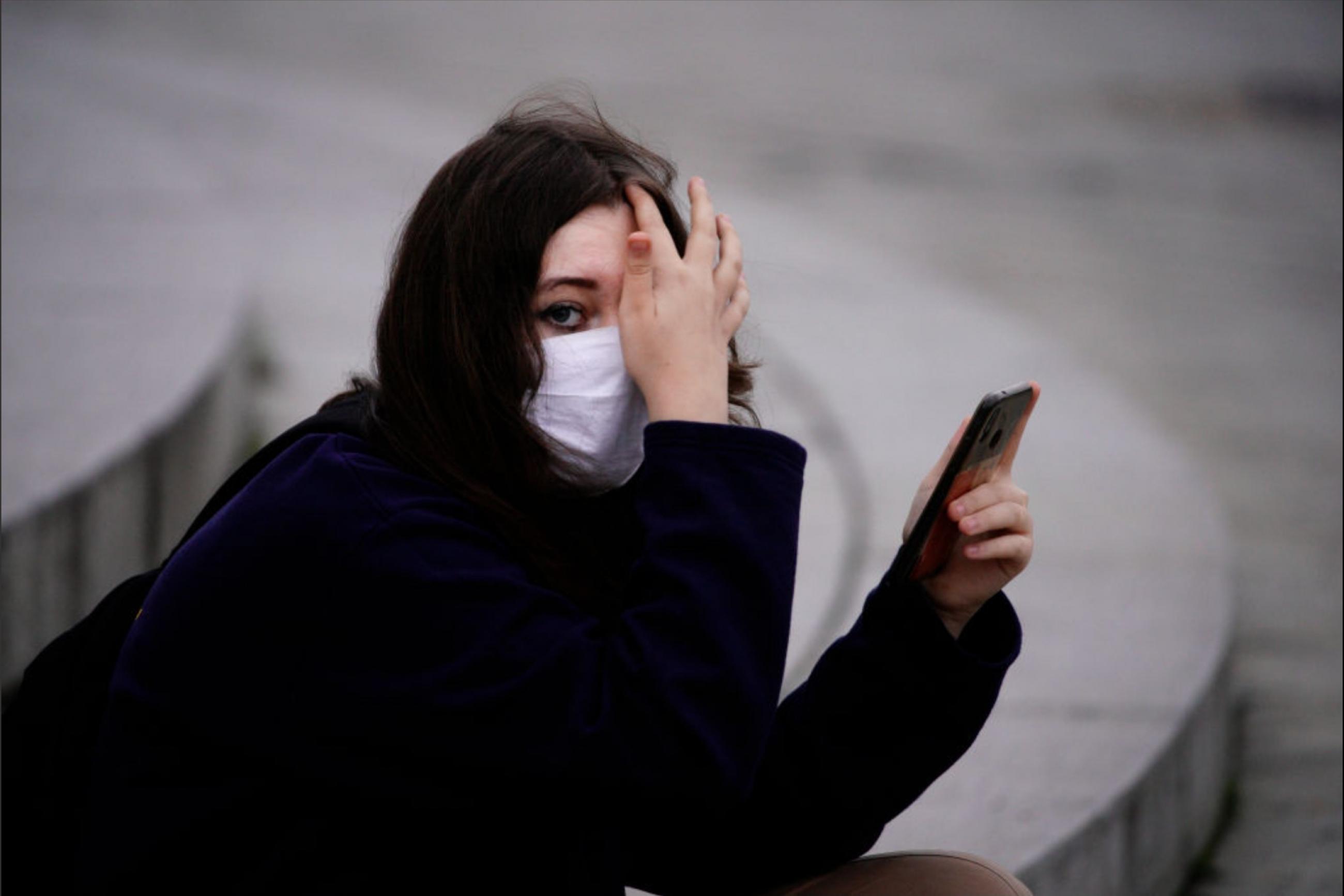 A young woman with dark hair in a black coat and white mask holds a black phone in one hand and covers part of her face with the other. She looks serious as she sits on grey stone steps in Warsaw, Poland.