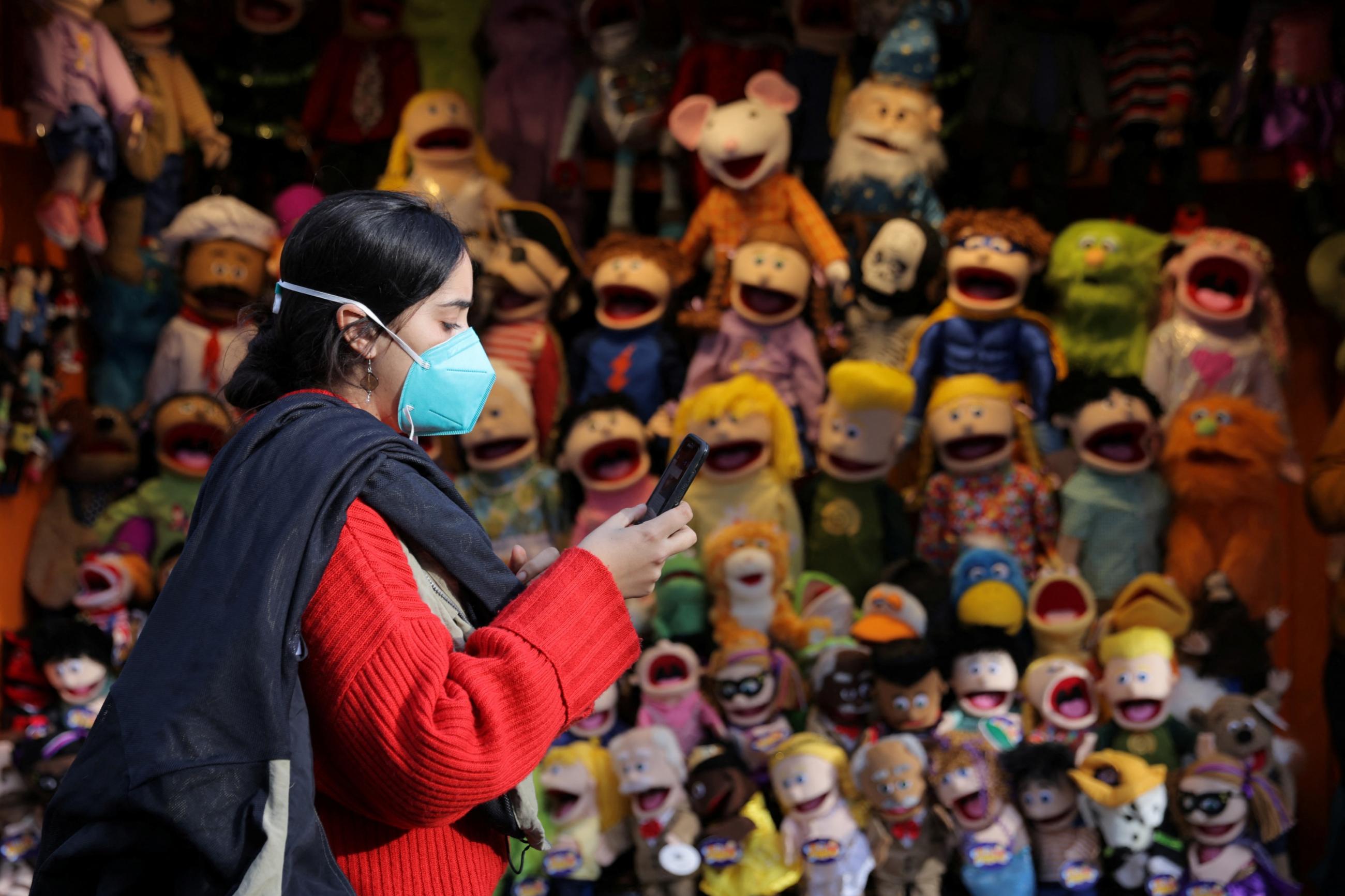 A woman in a red coat and blue mask checks her phone as she walks by a stall with colorful children's puppets
