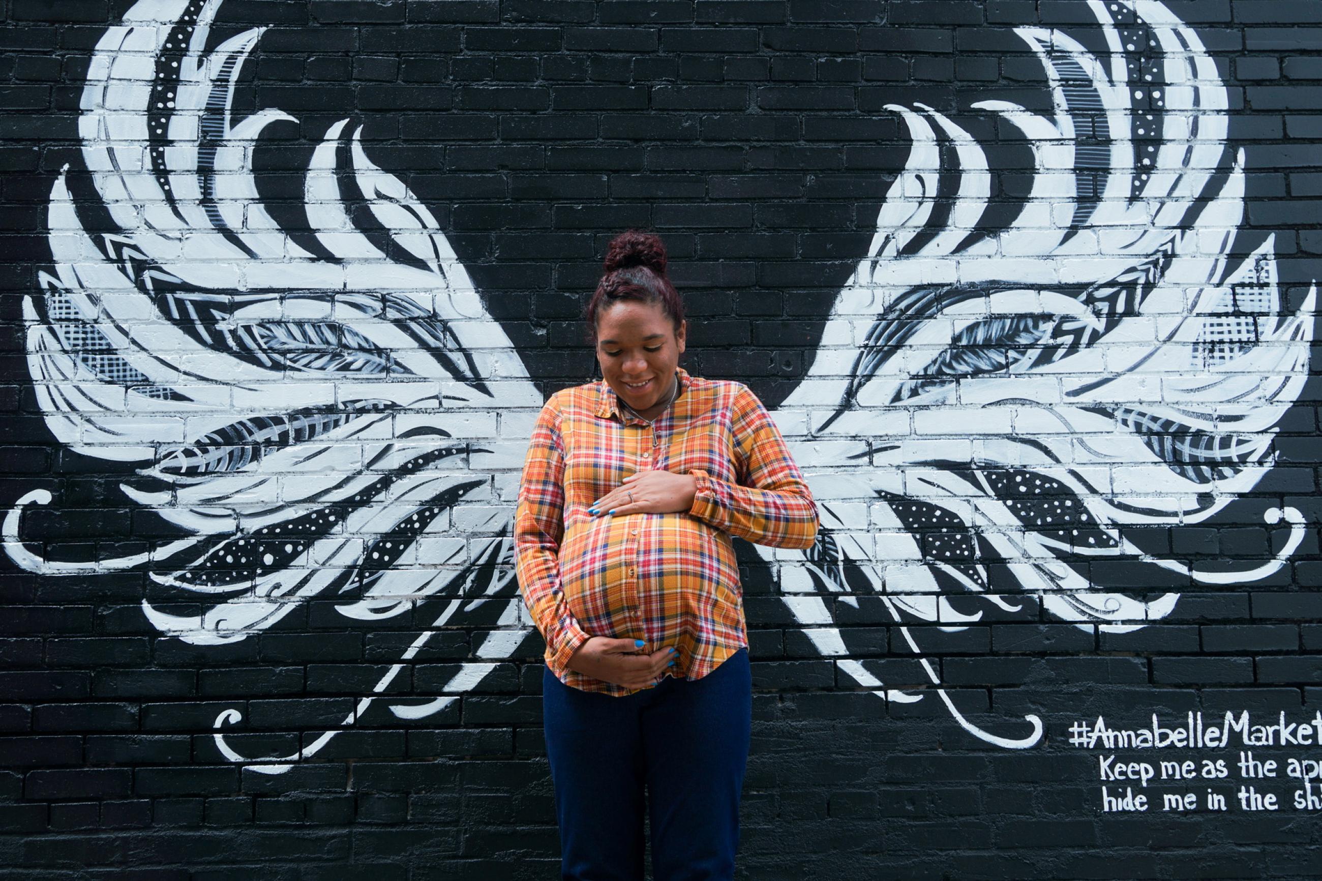 A Black mother in an orange shirt lovingly holds her pregnant belly in front of a mural on a black brick wall that gives her white angel wings in Anna, Illinois, U.S., May 10, 2021.