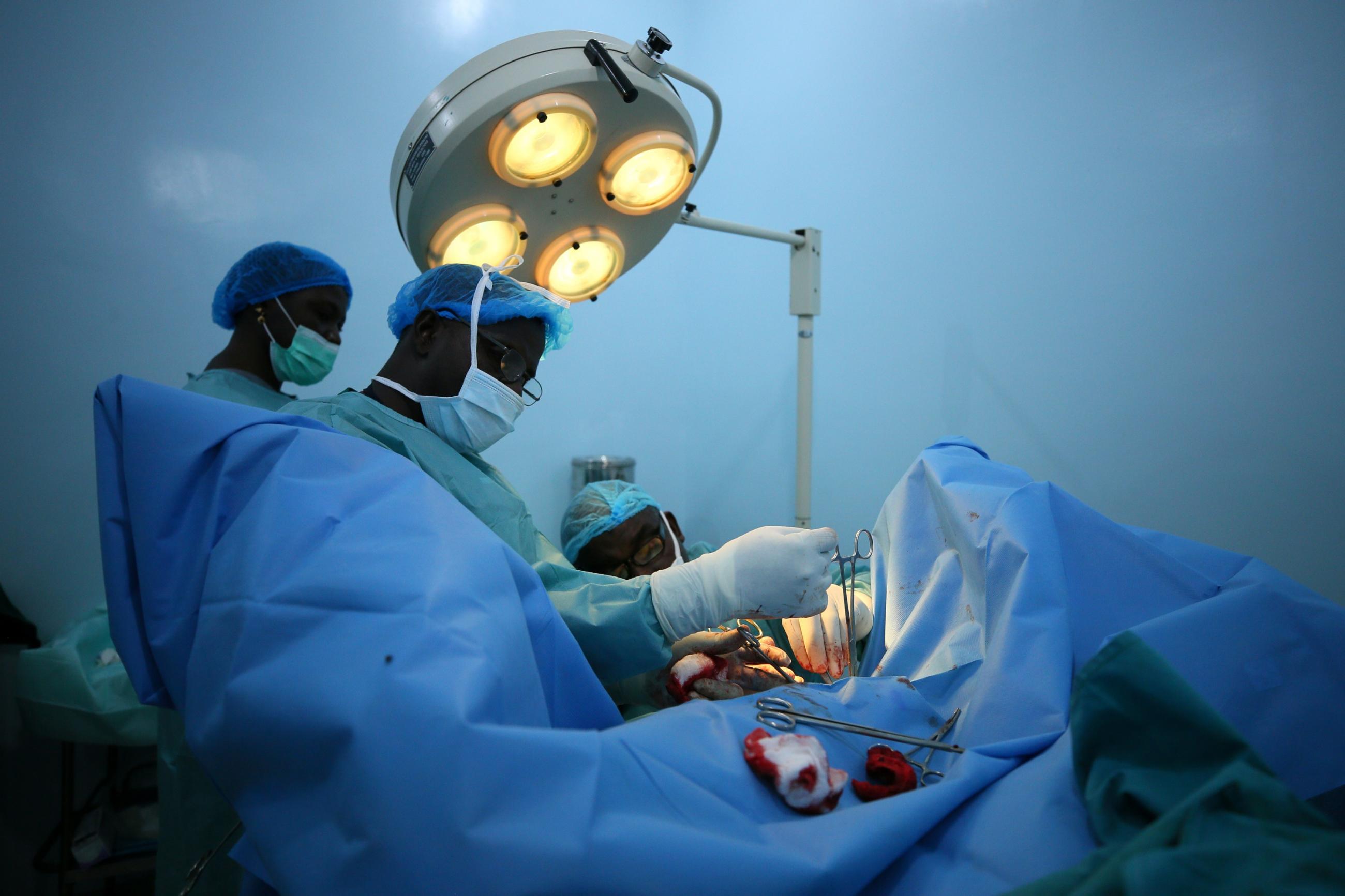 Nigerian surgeons in blue gowns and masks operate on a patient hidden beneath a blue sheet under a large surgical lamp in a room with blue walls. 