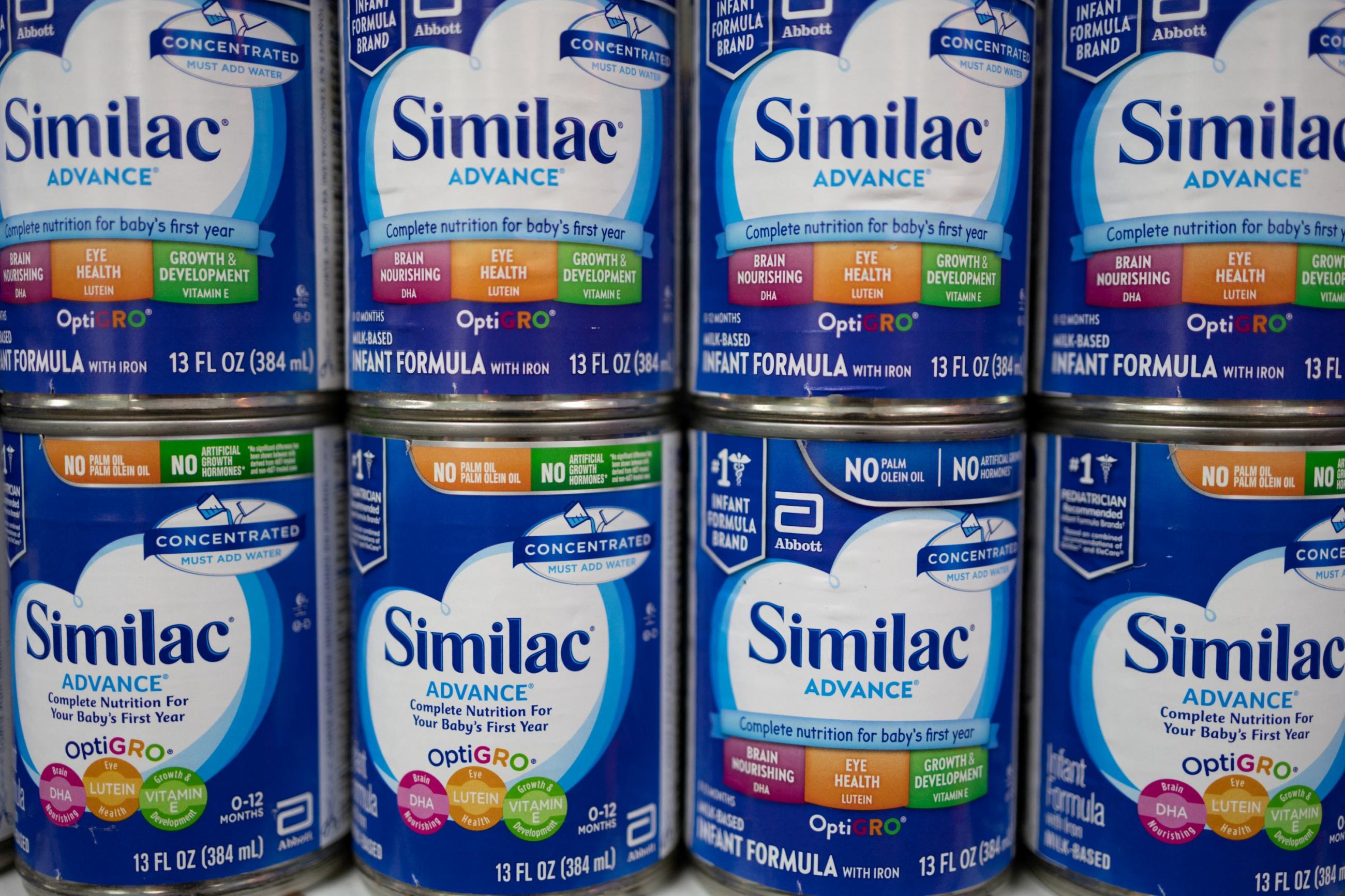Eight blue cans of baby formula with white, orange, and green labels that read "Similac" stand in stacks of two on a pharmacy shelf.