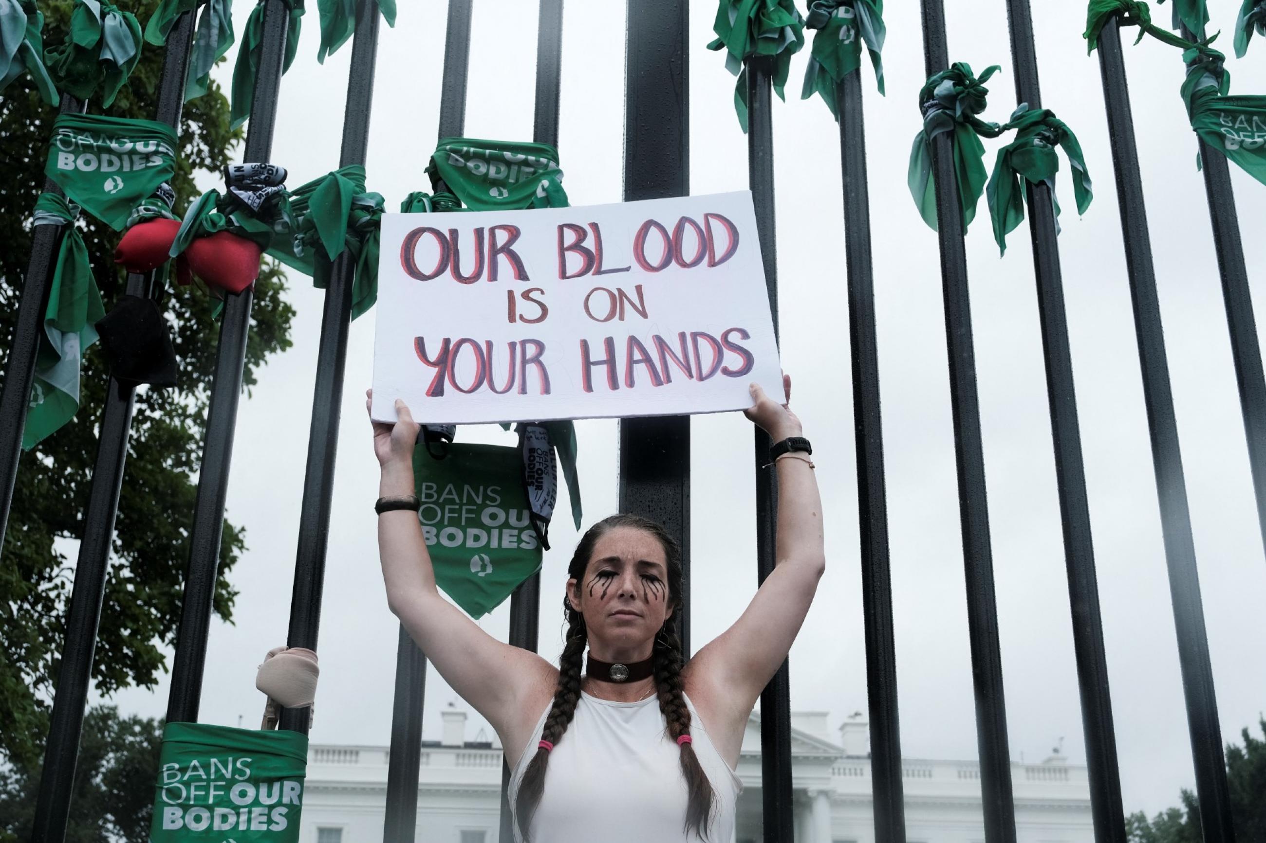 A woman with two braids wearing a white tank top holds up a sign that reads "our blood is on your hands" in front of the white house