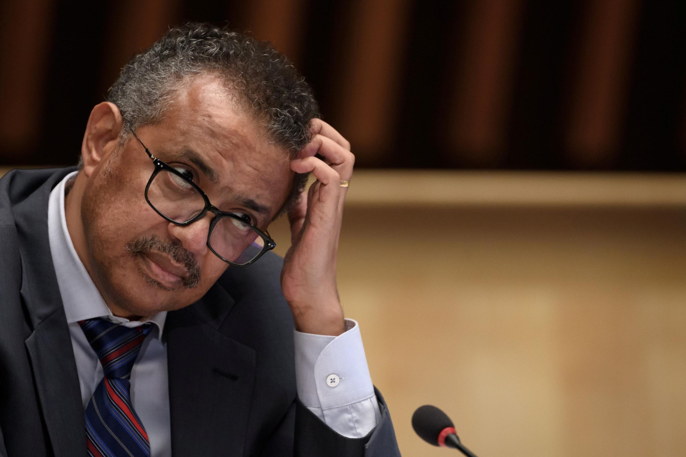 WHO Director-General Tedros Adhanom Ghebreyesus scratches his head with a quizzical expression while seated at a wooden podium 