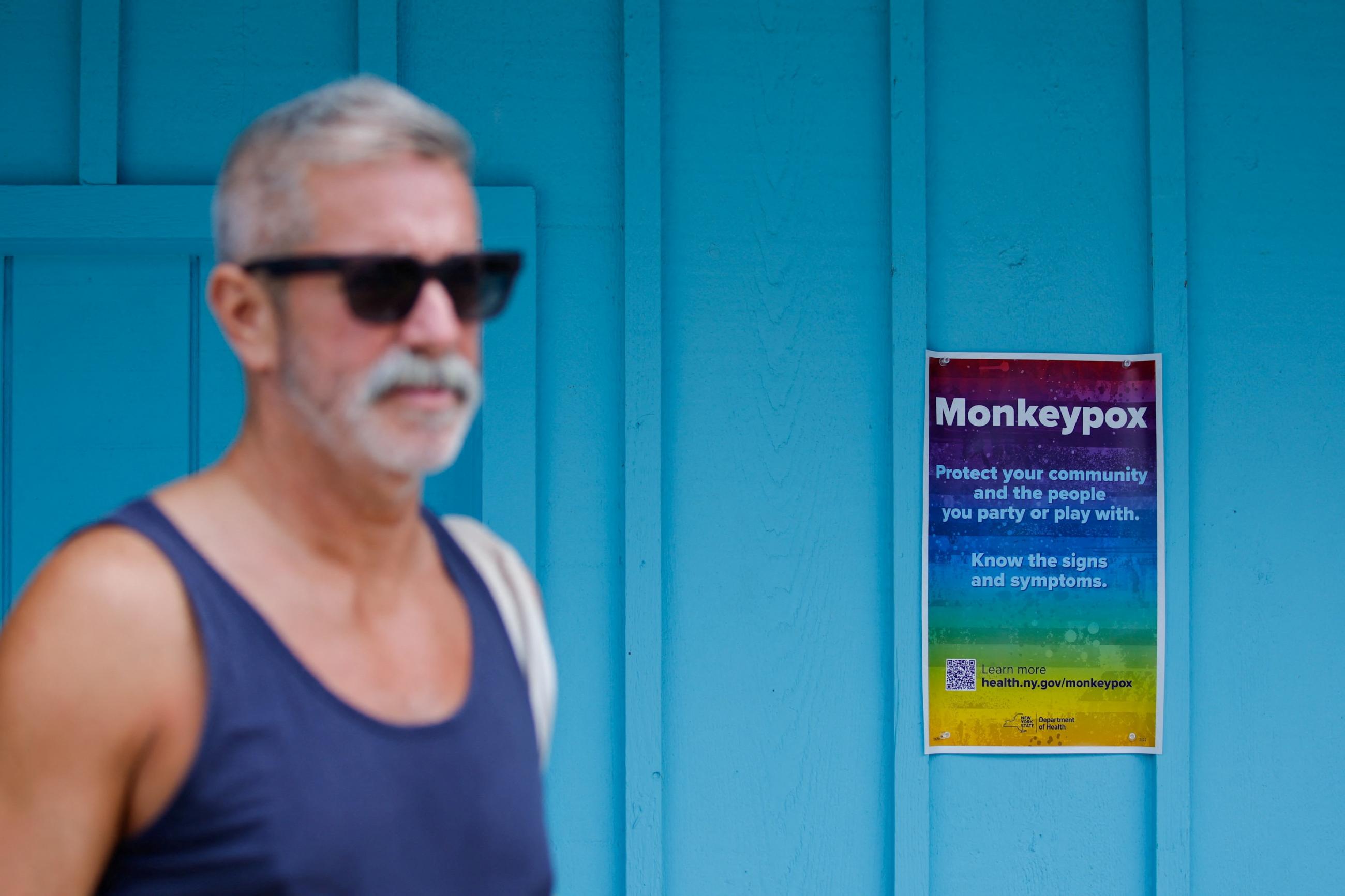 A grey-haired bearded man in a blue tank top and sunglasses walks past a turquoise wall with a rainbow poster that reads "Protect your community and the people you party or play with. Know the signs and symptoms" placed there by the New York Department of Health