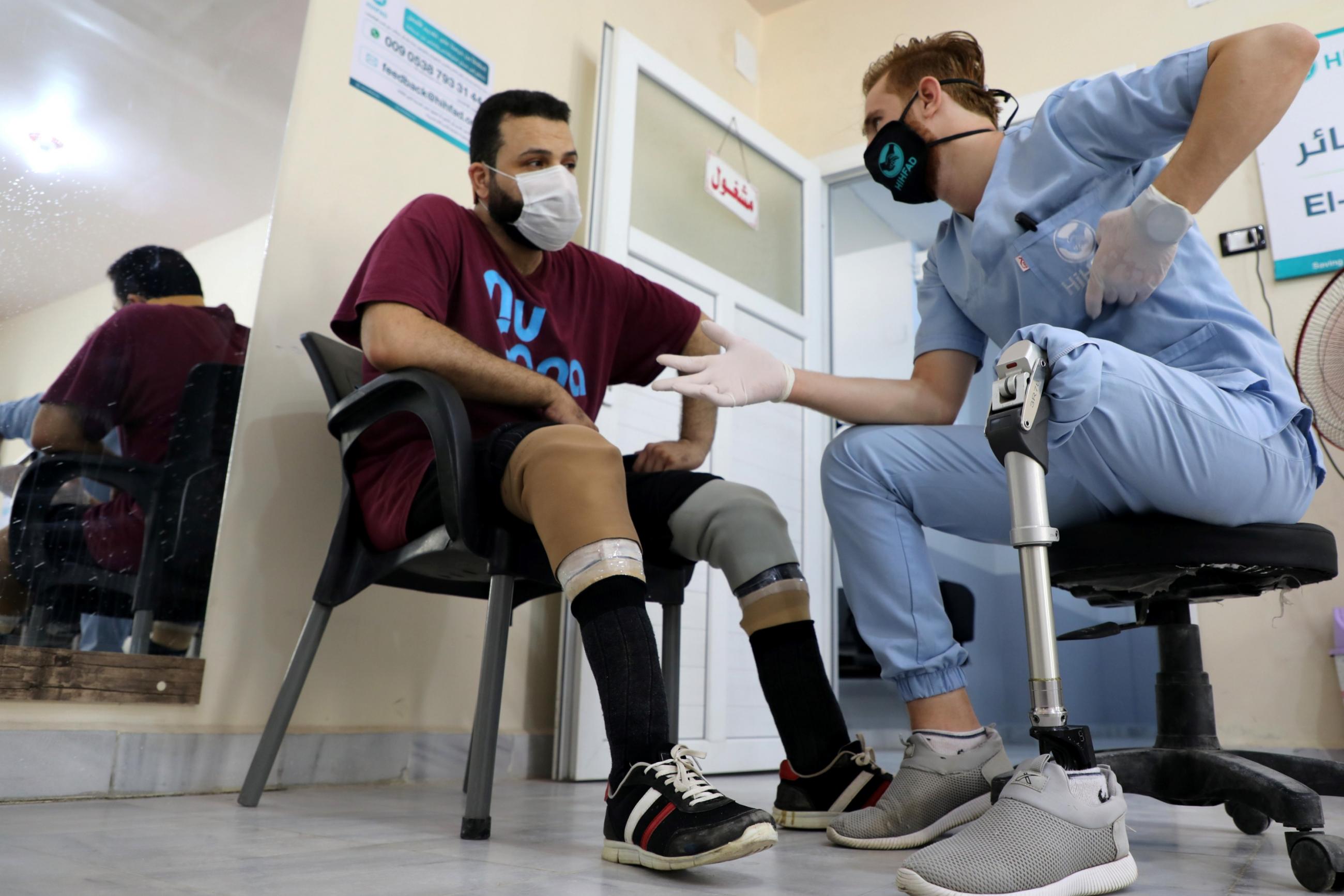 A Syrian physical therapist with a prosthetic leg, wearing blue scrubs and gesturing compassionately talks to a patient, who also has a prosthetic leg at the Al-Bab Center for prosthetics. Both men are sitting in chairs talking eye-to-eye. 