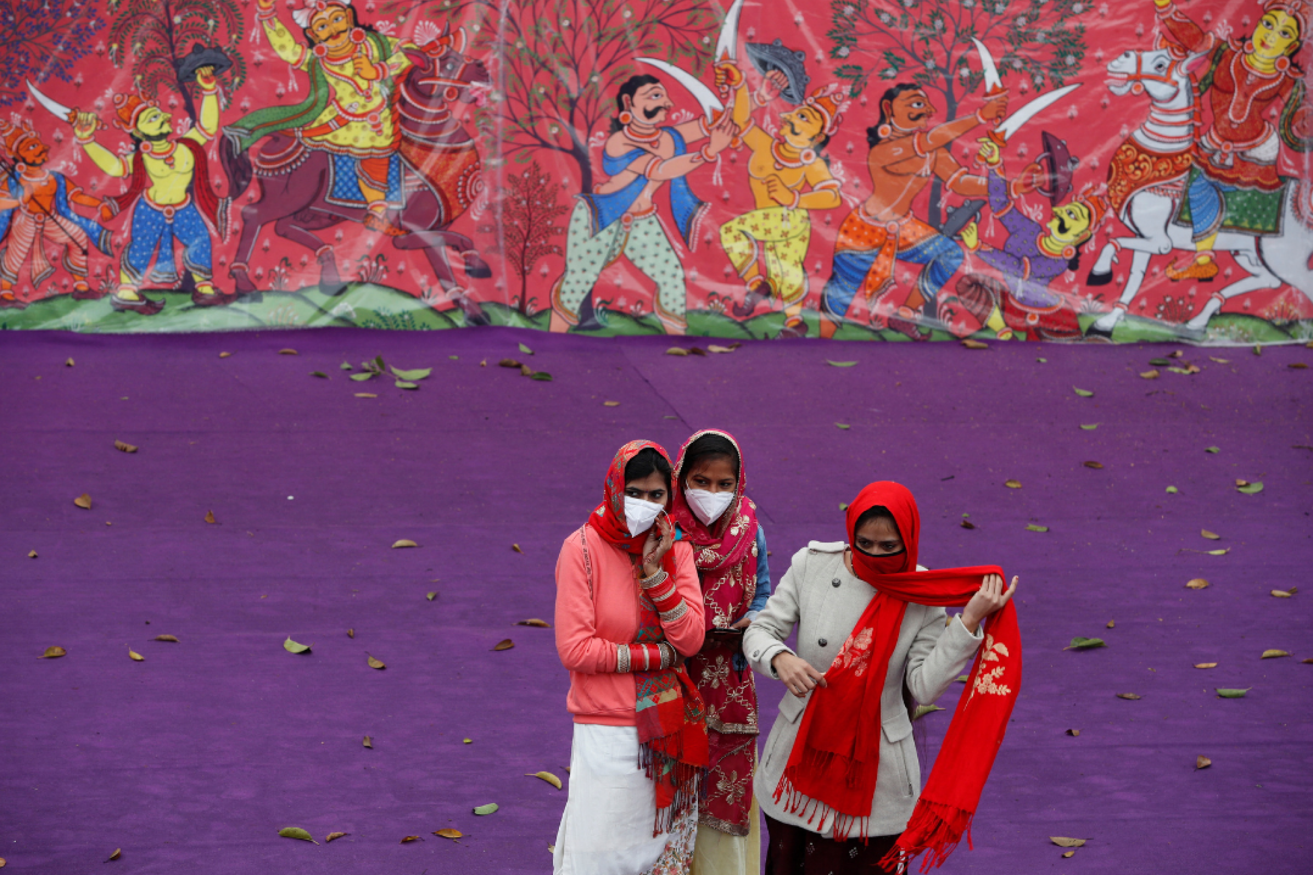 Women wearing protective face masks and colorful red and white saris stand in front of a purple wall and a color mural as they watch a dress rehearsal for the Republic Day parade, in New Delhi, India, on January 23, 2022. REUTERS/Adnan Abidi