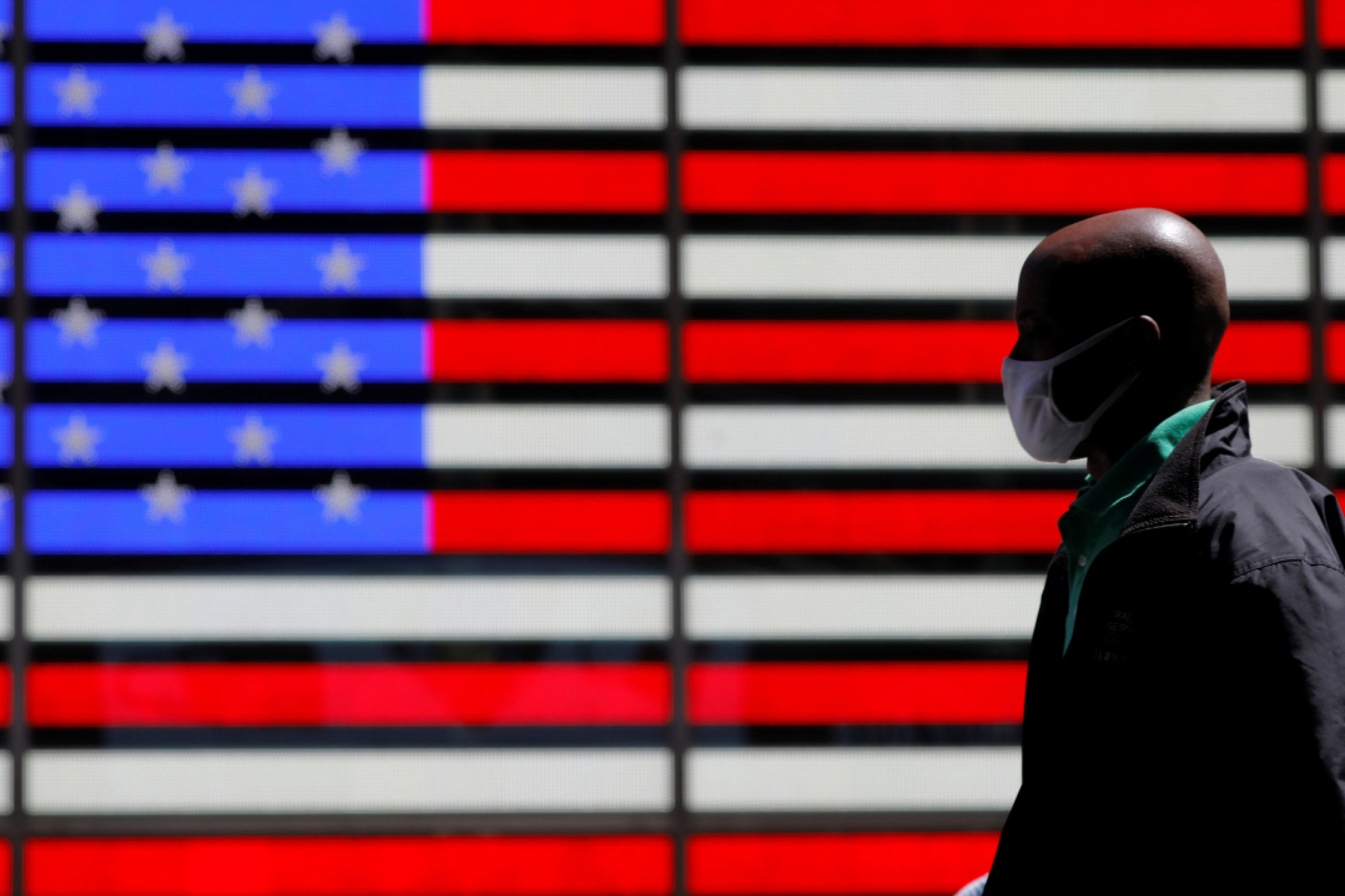 A giant red, white, and blue lit-up U.S. flag can be seen behind a person wearing a mask while walking in Times Square after the CDC announced new guidelines regarding outdoor mask wearing and vaccination during the COVID outbreak in New York City, New York, on April 27, 2021. 