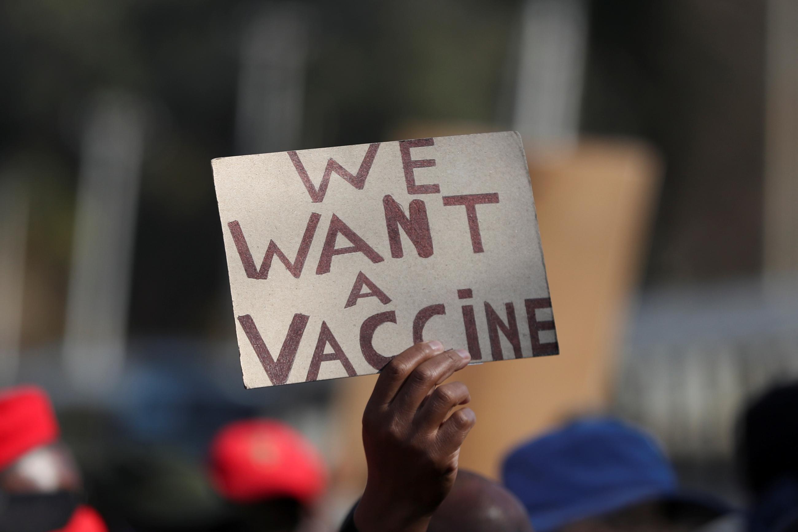 Hand holding up a sign that reads "We Want a Vaccine"