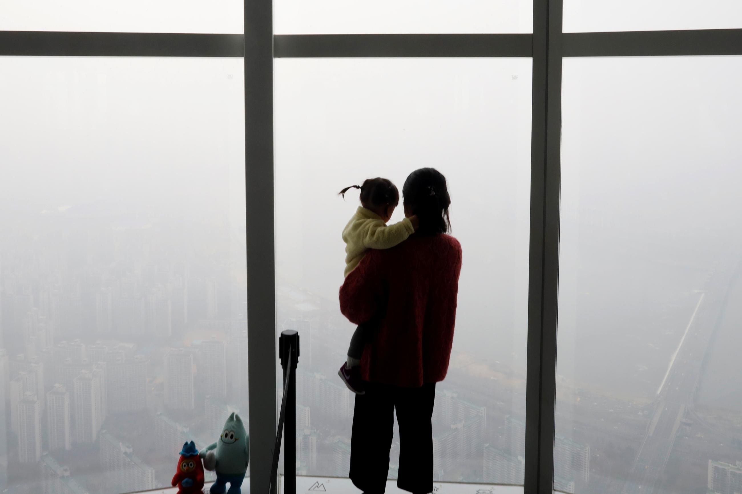 A woman holding her baby in her arms looks out a window at the city shrouded by a fine dust during a polluted day.