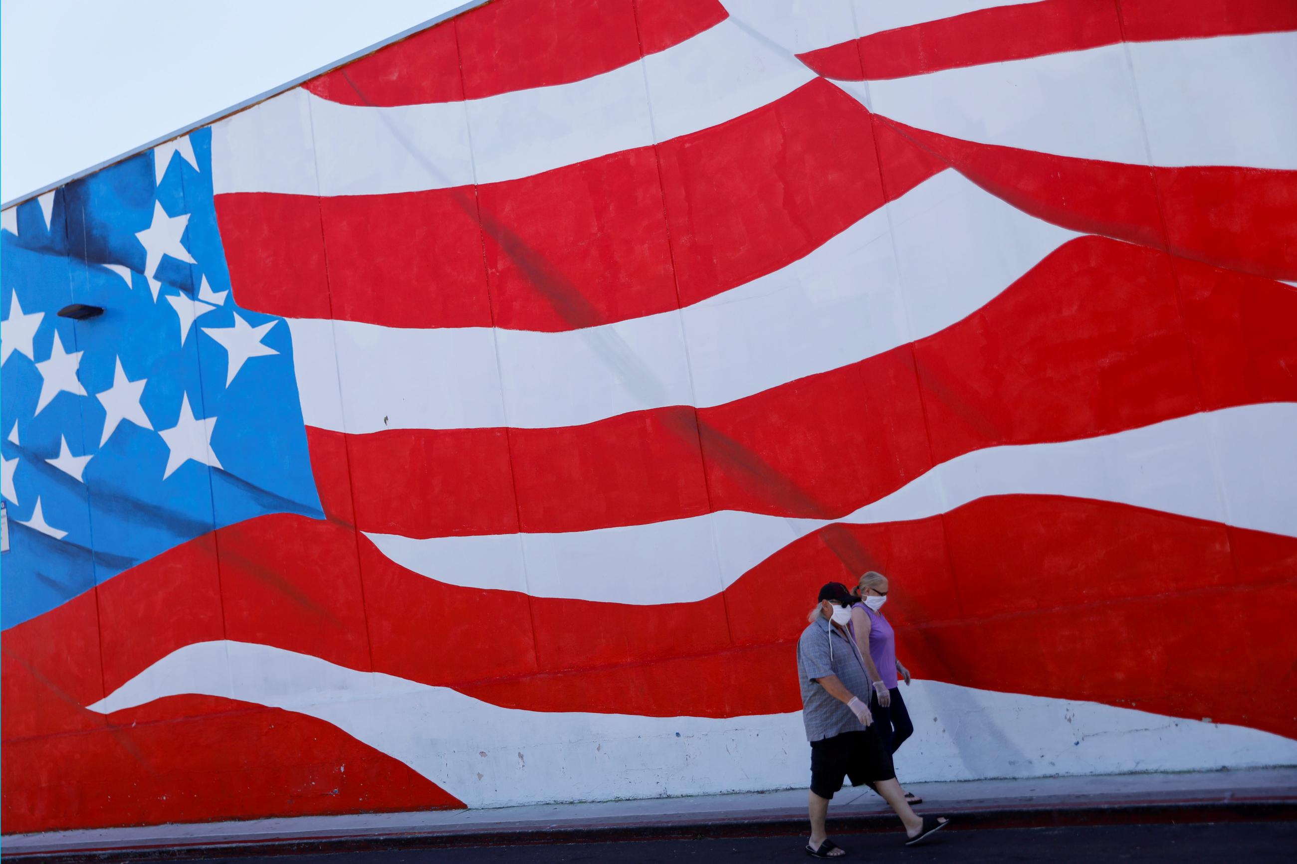 People wearing masks walk past an American flag painted on a building during the outbreak of COVID-19.