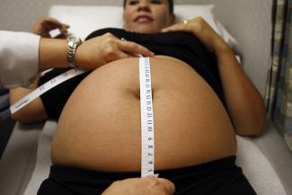 A woman receives a prenatal exam at the Maternity Outreach Mobile in Phoenix, Arizona. The picture shows a woman lying back on a hospital-type bed with her shirt pulled up exposing her late-term belly. The hands of a health care provider stretch a tape measure vertically across her belly.