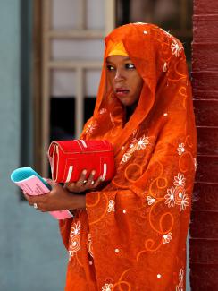 A woman waits at a polio immunization health center in Maiduguri, Borno State, Nigeria, on August 29, 2016. There is striking color in this photo. She has a bright orange scarf around her head and shoulders and leans against a brick wall that is painted dark red. She holds pink and blue copies of papers in her hands, and the light blue wall of a building can be seen in the background. She holds a bright red bag. The look on her face is serious.