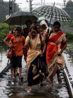 Several women and at least one boy are walking in a group. One of them is talking on a cell phone. Several are sharing umbrellas. The water, even on the train tracks, which are presumably slightly elevated, is up to their ankles. 