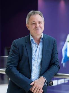 The picture shows Jeremy Farrar wearing a blue shirt and blazer, smiling as he leans on sleek railing and standing out against a highly bokeh background. 