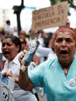 Health care workers protesting shortage of medical supplies outside a hospital in Caracas, Venezuela on November 19, 2019. The picture shows an older woman in the foreground wearing blue hospital scrubs shouting and looking slightly off camera. She is surrounded by other protestors, many of whom are waving protest signs and also shouting. REUTERS/Carlos Garcia Rawlins