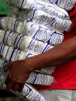 Picture shows the torso of what appears to be a young man heavily burdened with more than a dozen large, one-pound bags of salt. He wears a red shirt and walks against a green backdrop and the bottom bag appears to be slipping out of his grasp. 