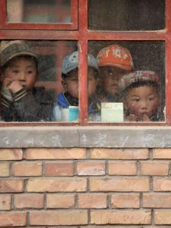 We see the adorable faces of several small children looking out weather-beaten window panes hung on a yellow brick wall. Faded chalk writing of chinese characters can be seen scribbled on the concrete sill.