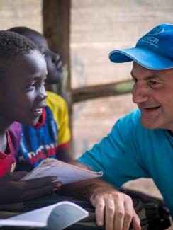 Picture shows Dr. Salama wearing a blue hat talking to several boys in a classroom. They are all smiling. 