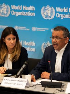 The photo is taken of a press conference with three people at a table in front of microphones. They look very serious. 