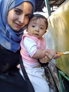 The picture shows a closeup of mother and child, both looking at the camera. She wears a blue had scarf. The baby has a pink pullover and holds a brush and is slathering yellow-green paint on the metal shell of a vehicle.