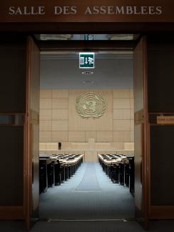 The picture shows a stately entrance to a hall with the words “SALLE DES ASSEMBLEES” in big block letters over the door. The doors are open revealing a wide aisle to the stage which has a massive UN logo hanging over it. 