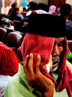 The picture shows a crowded waiting room with a man in the foreground with a red cloth draped over his head and holding what appears to be a bag of ice against his cheek. He looks like he is suffering. 
