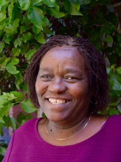 The photo shows Edinah smiling in front of a background of leafy brush.