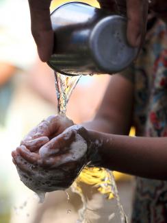The picture is a close-up of the girl’s hands, showing the water droplets cascading of her soapy hands. An adult pours the water from a metal cup. 