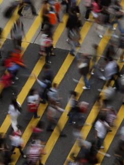 Picture shows the street from above, a wide pedestrian walkway with broad yellow lines painted at an angle. Dozens of people can be seen in the frame, all blurred due to fast movement and a slow shutter. 