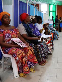 Image shows a row of several women wearing colorful scarves on their heads. The camera is looking down the row, and the woman in the foreground is looking at the camera. Two women in the background appear to be talking. 