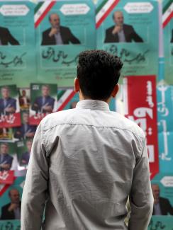 The photo shows the man standing in front of a board that is placarded with multiple political campaign posters, many of them identical and pasted over and over. The predominant colors are green, white, and red—like the Iranian flag. 