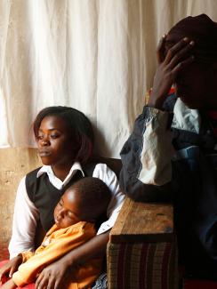 Picture shows a young woman holding a child looking off to the left while another woman, obscured by shadows, sits next to het with her head resting against her hand, as if in anguish. 