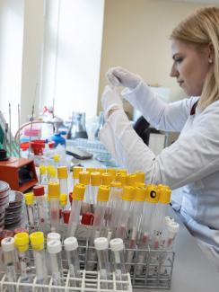 Picture shows a lab worker in a white lab coat sitting at a bench surrounded by test tubes preparing samples for analysis. 