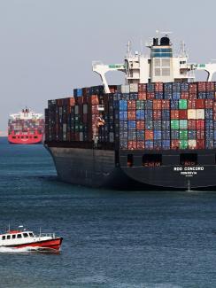 The picture places the viewer in the wake of a massive freighter stacked high with metal shipping containers as a smaller patrol-type boat passes in the foreground. 