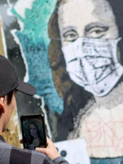 The photo shows a man with dark hair and a black cap taking a cell phone photo of a likeness of Da Vinci's most famous painting with Mona wearing a mask. 