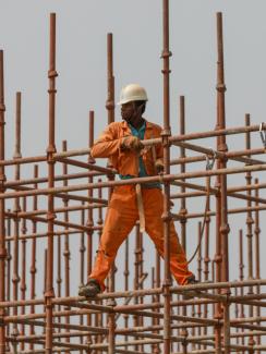Picture shows a man in a bright orange jump suit and a yellow contruction hat balancing with his legs wide apart on a metal scaffolding against a grey sky. 