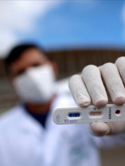 The photo shows a health worker holding a test at the camera with a stadium in the background. the photographer is using a telephoto/macro lens so that everything is blurred out in the shot, including the man, with the exception of the small plastic cassette-like test.  R