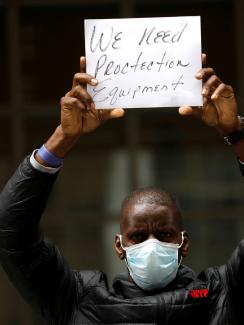 The image shows a man wearing a mask and holding a small sign above his head that says, "We need protection equipment." 