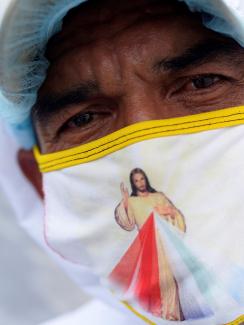 The photo shows the worker in protective head covering and mask. on his mask is an image of Jesus with a rainbow colors on his robe. 