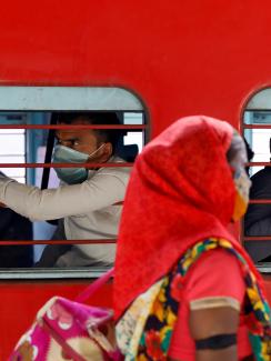 The image shows a bright red train with people inside and a woman walking by the train outside. 