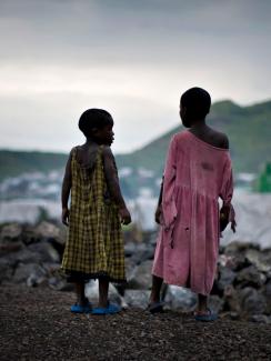 The photo shows two girls standing on a hill overlooking a camp filled with white tents at sunset. 