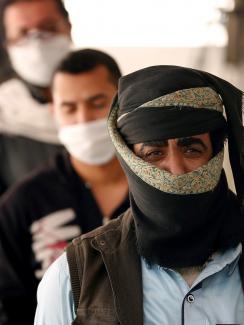 The photo shows several men lined up in a queue. They are all wearing facemasks. 