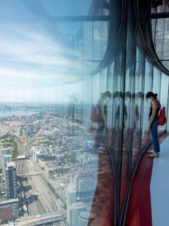 The photo shows a woman wearing a mask looking out from the skyview of the tall building with the Toronto cityscape beneath her. 