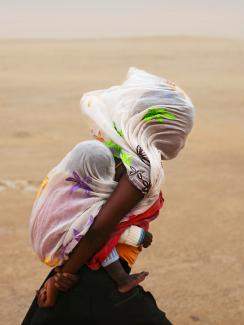 The photo shows a woman with a baby on her back walking through a desert. The wind is blowing strongly, as evidenced by the billowing cloth on their heads. 