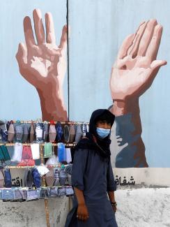 The photo shows a man in front of a small cart with masks on display. in the background is a mural of hands raised. 