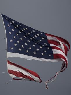 The photo shows a torn U.S. flag blowing against a dark grey sky. 