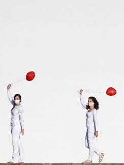 The photo shows four artists dressed in white walking in front of a massive white backdrop. Each holds a red balloon. 